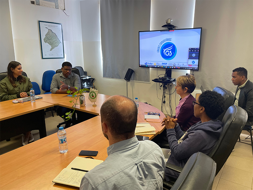 Attendees of visiting sitting around tables having a meeting in a room, with computer monitor in background.