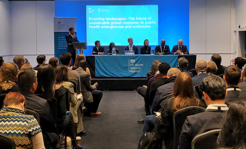 Six panelists sitting on stage infront of projected screen at UKHSA conference 2025. Photo from back of room showing seated audience. 