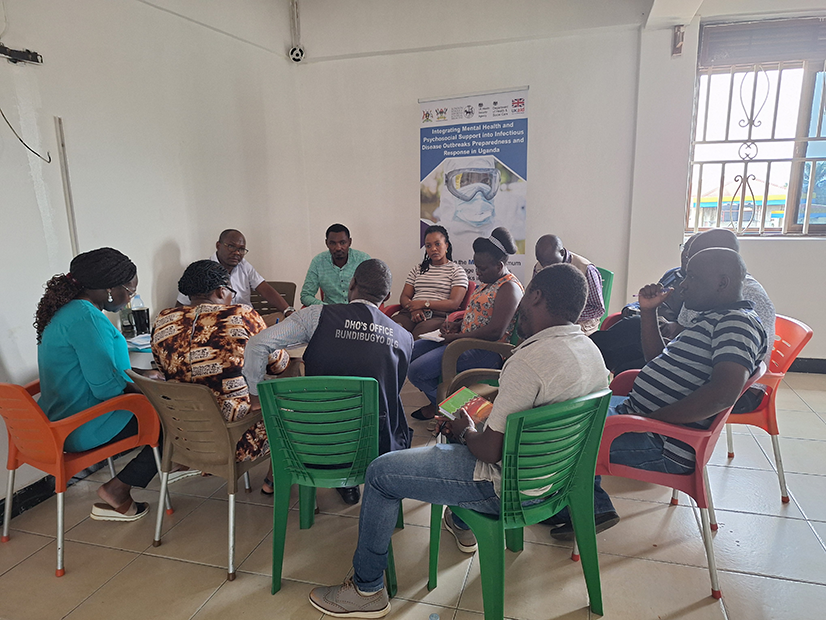 MHPSS Uganda workshop participants sitting together in circle on plastic chairs with workshop banner in background of classroom.
