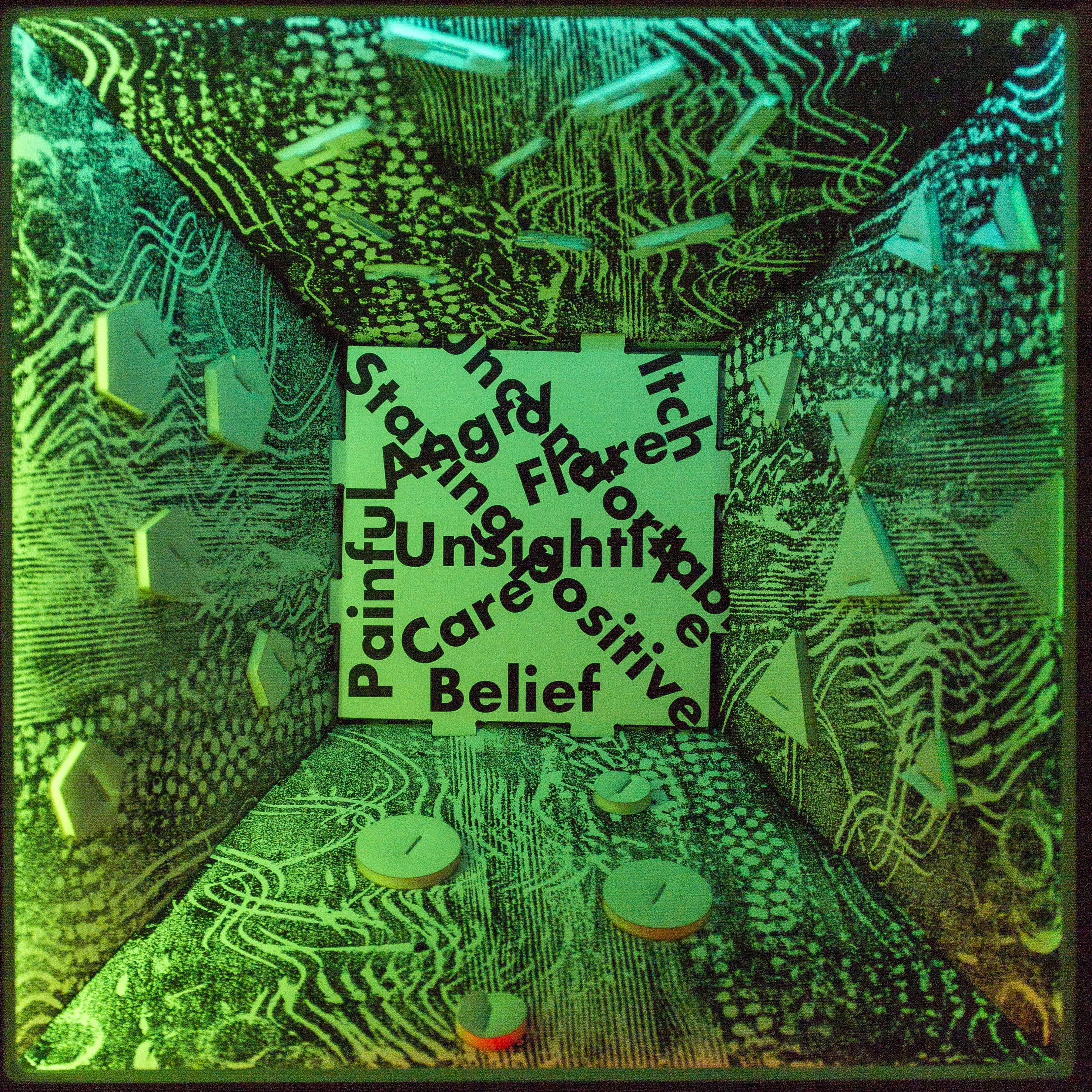 Hollow box lit by LED lights which are mostly green and light blue colours. Patterns and shapes are on the sides of the box and in the middle are a selection of words including: painful, itch, belief, staying positive, angry, and uncomfortable.