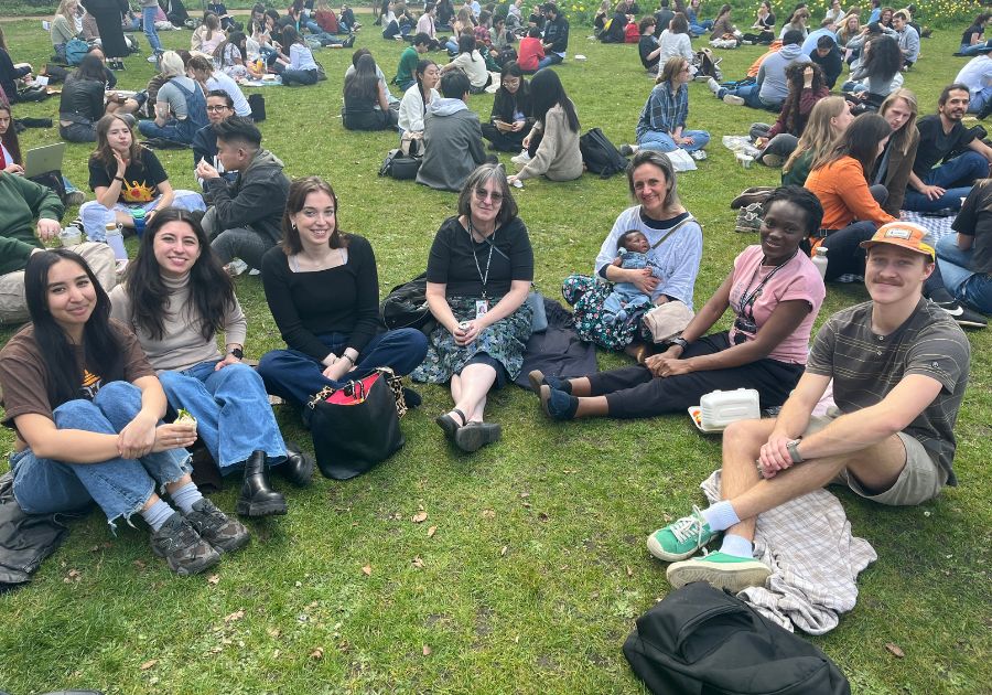 Students and Programme Director from the MSc Demography and Health sit outside in Gordon Square park and smile towards camera surrounded by lots of groups socialising and having picnics.