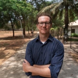 Photo of a white man with blonde/brown short hair and glasses wearing a blue striped shirt with trees in the background