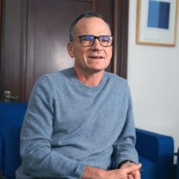 Photo of a white man with grey/black hair and glasses, wearing a blue jumper