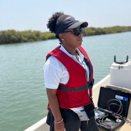 Black woman wearing a cap and sunglasses standing on a boat with a red life jacket on