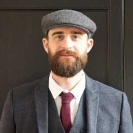 White man with beard and hat wearing grey suit and red tie