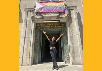 Student, Alexandra, stands outside LSHTM underneath an LSHTM Progress Pride flag with her hands in the air.