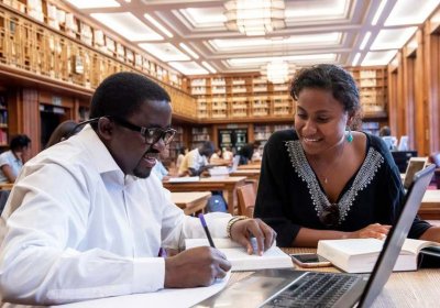 Two individuals sitting in the LSHTM library are looking over pieces of work