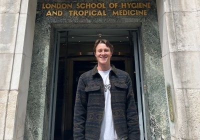 Man stands smiling outside LSHTM building with dark jacket and white t-shirt.
