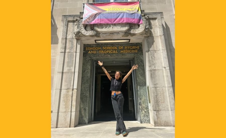 Student, Alexandra, stands outside LSHTM underneath an LSHTM Progress Pride flag with her hands in the air.